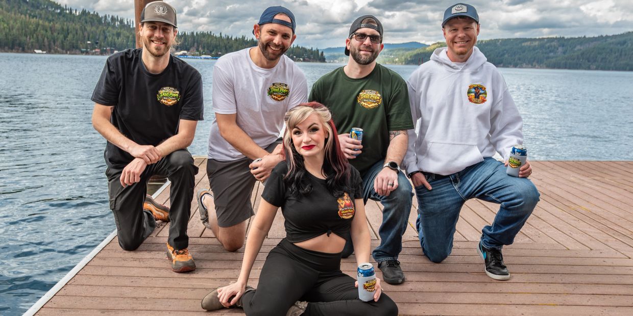 Group of five young adults posing on a dock by a lake, smiling and holding drinks.