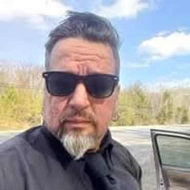 Man wearing sunglasses and black shirt outdoors near a car.