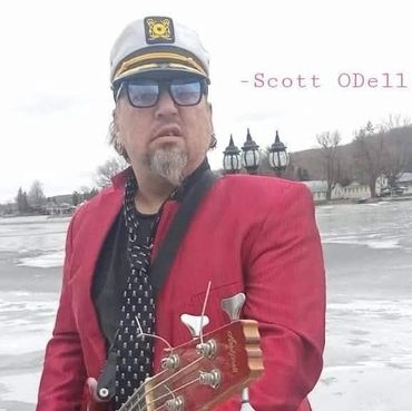 Man in captain's hat and red jacket holding a guitar by a frozen lake.