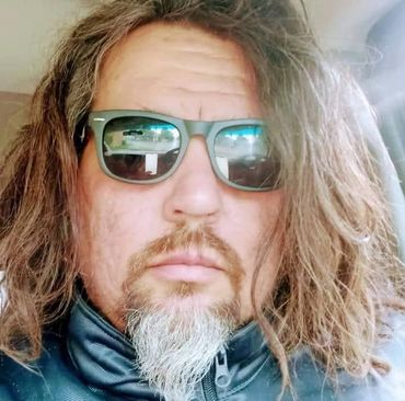 Man with long hair, beard, and sunglasses inside a car.
