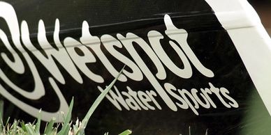 A black carbon surf ski kayak with a Wetspot Watersports sticker sits on the grass awaiting repairs.