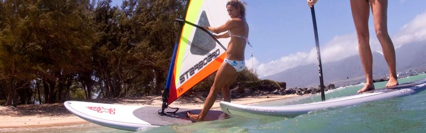 A fit woman windsurfs past another on a Stand Up Paddleboard.