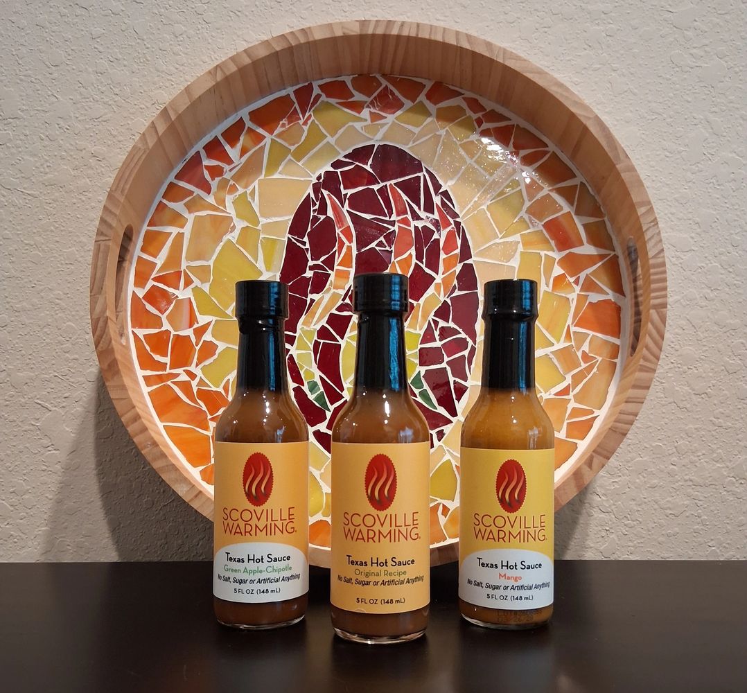 Three bottles of Scoville Warming brand hot sauce in front of a colorful round mosaic tray.