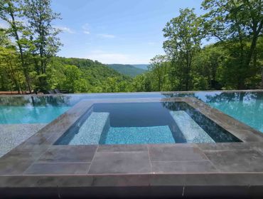 Infinity pool with hot tub overlooking lush green mountains under a clear blue sky.