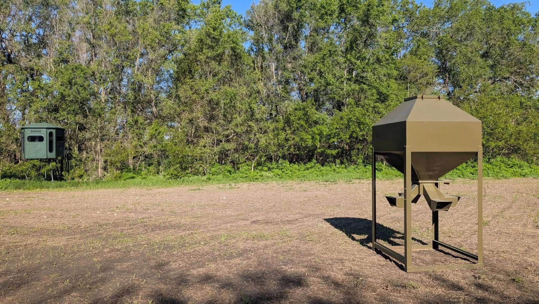 A hunting blind and a green feeder in a clearing near trees.