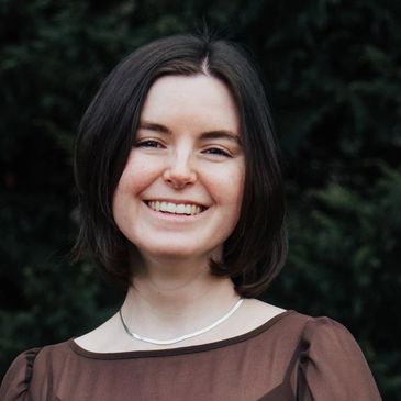Smiling woman with short dark hair in a brown top against a dark leafy background.