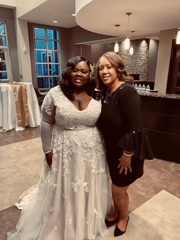 Two women posing indoors, one in a white wedding gown and the other in a black dress.