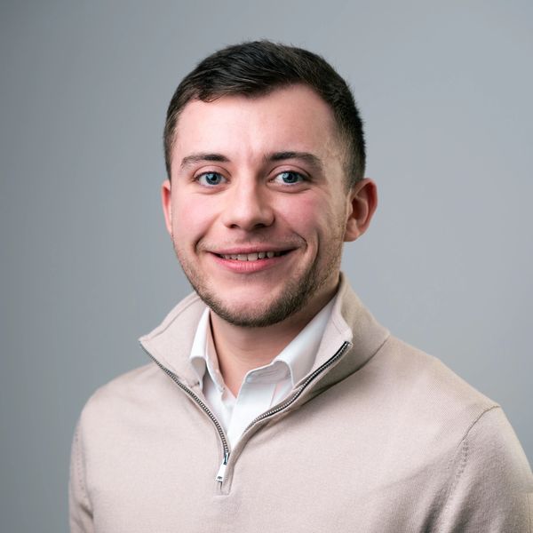 Young man smiling in beige sweater against gray background.