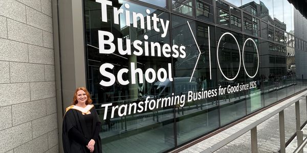Graduate in cap and gown stands outside Trinity Business School celebrating 100 years.