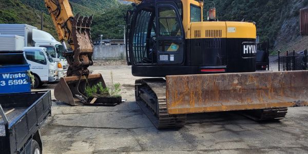 Yellow Hyundai excavator parked on a concrete surface near trucks and greenery.