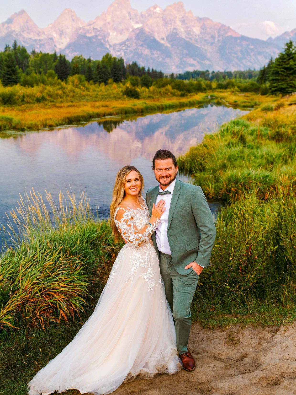 A newlywed couple poses by a serene river with mountains in the background.