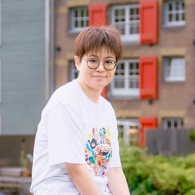 Person with glasses and short hair smiling outdoors near a building with red shutters.
