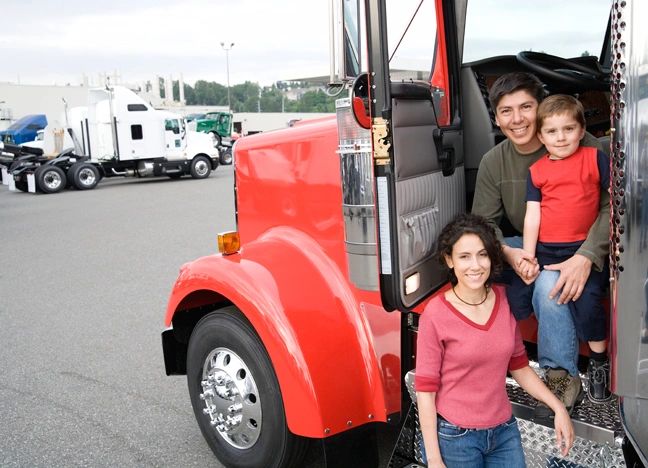 Family posing happily by a red semi-truck on a parking lot.