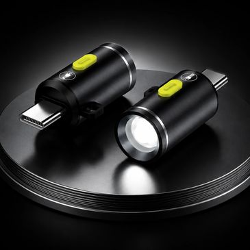 Two compact black USB-C LED flashlights with yellow buttons on a reflective black surface.