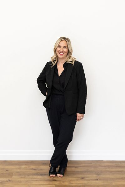 Smiling woman in black business attire standing against a white wall.