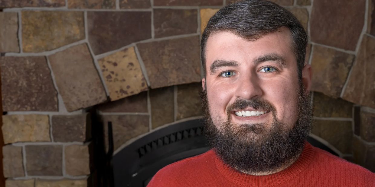 Smiling man with a beard in a red sweater against a stone wall background.