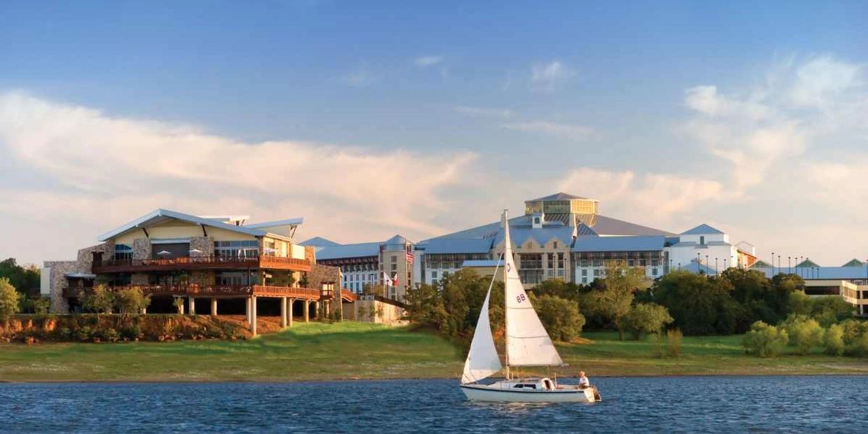 Sailboat glides on water near a lakeside resort under a blue sky.