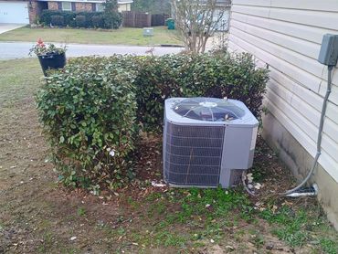 Outdoor air conditioning unit surrounded by trimmed bushes near a house.