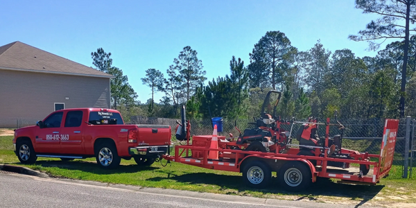 Red pickup truck towing a red trailer with lawn care equipment on a sunny day.