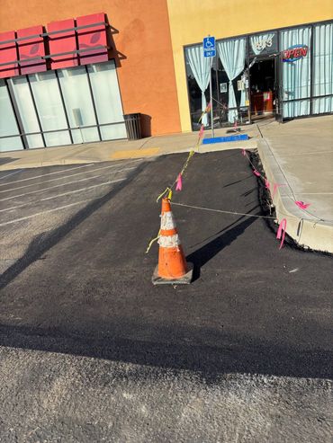 Freshly paved asphalt area alongside a commercial building with orange safety cones.