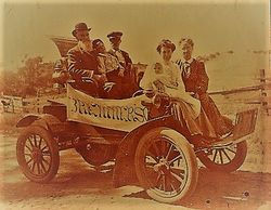 Bremner family in the early 1900s riding a horseless carriage with a banner advertising "Bremners."