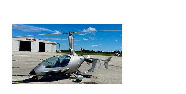 Silver gyrocopter parked on an airfield under a blue sky.