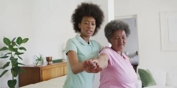 A young woman helps an elderly woman with arm exercises indoors.
