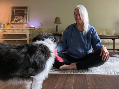 Woman sitting cross-legged on floor, playing with a dog indoors while trying to do yoga