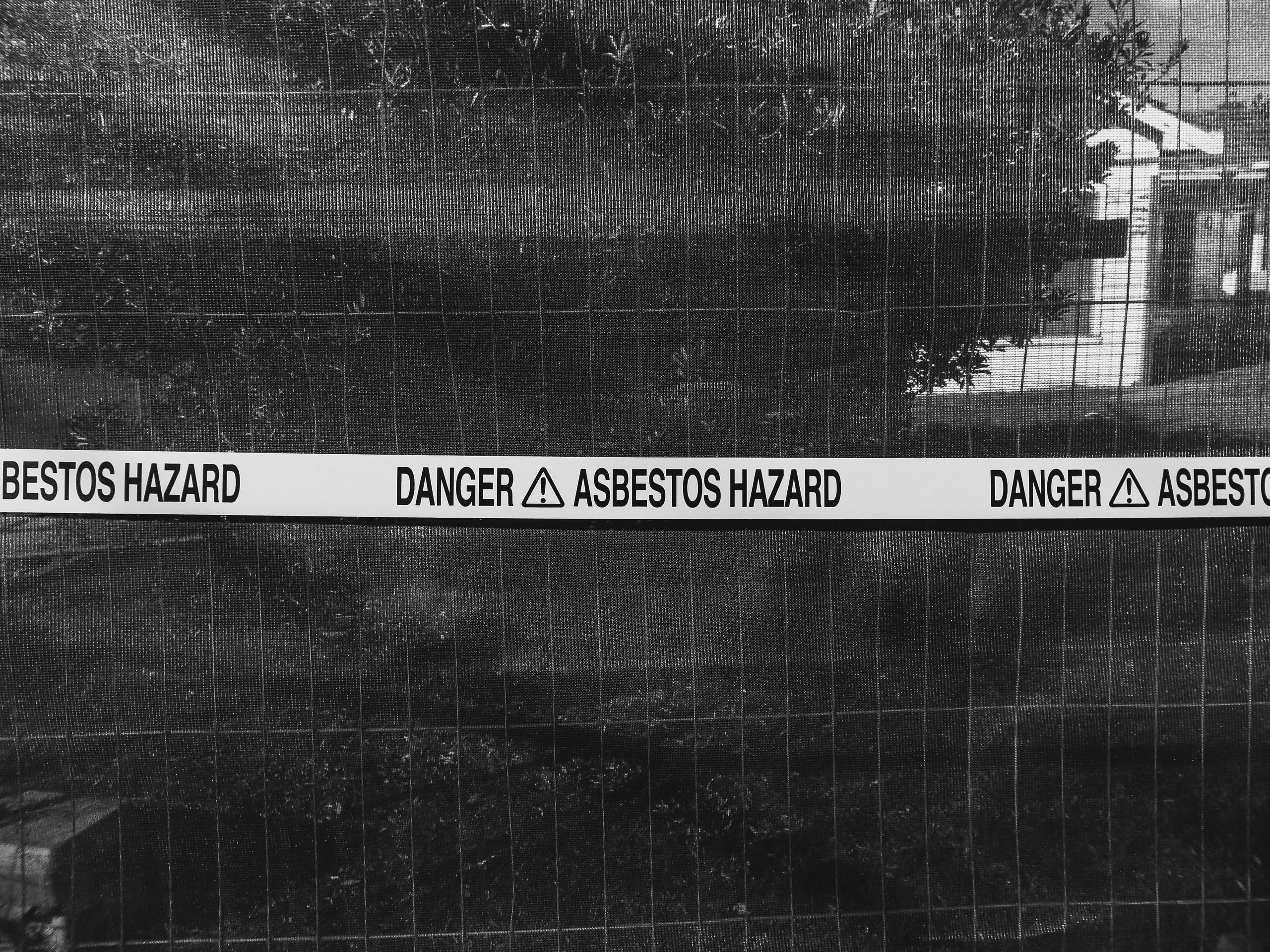 A wire fence with "Danger Asbestos Hazard" warning tape across it.