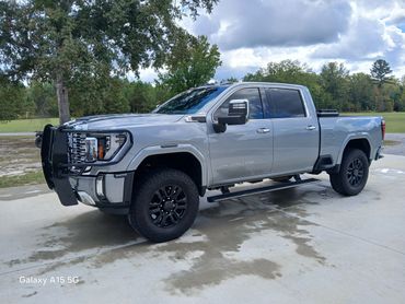 Silver GMC Denali HD truck parked on a driveway with trees and cloudy sky.