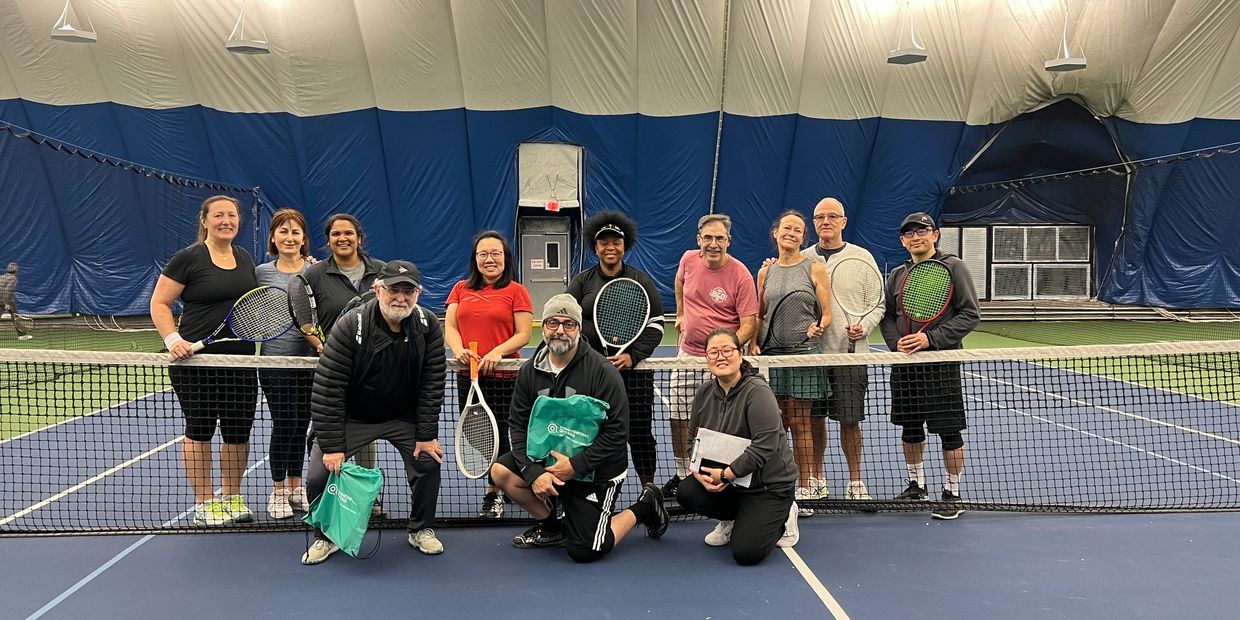 Group photo of tennis players posing on an indoor tennis court behind the net.
