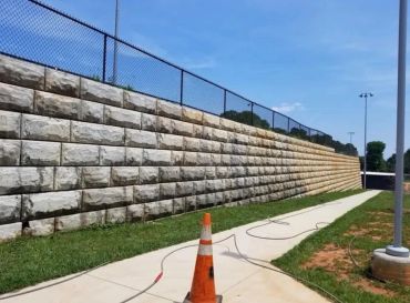 Stone retaining wall plus sidewalk with orange cone and fence.