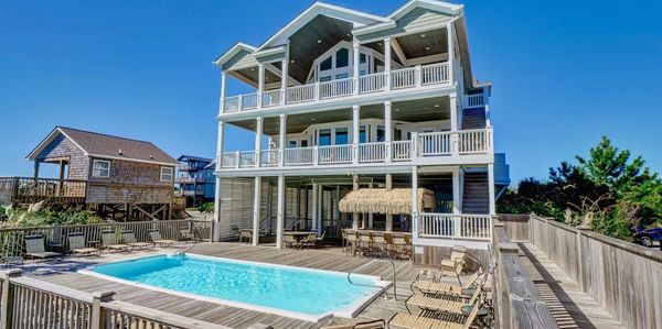 Modern three-story beach house with a pool and deck seating under clear blue skies.