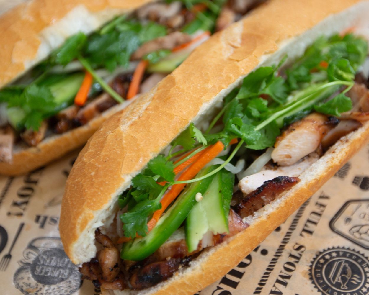 Two Vietnamese Banh Mi sandwiches with grilled meat and fresh herbs.