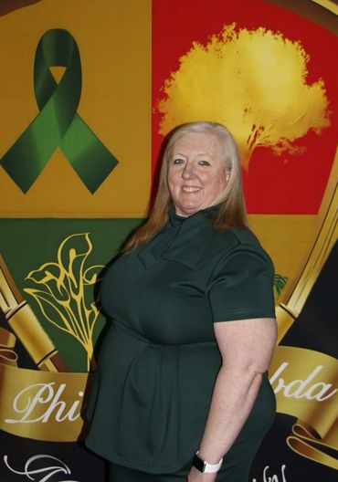 A woman in green stands smiling in front of a colorful Phi Beta Sigma banner.
