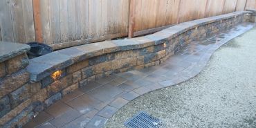 Stone retaining wall with embedded lights and a paved walkway beside a wooden fence.