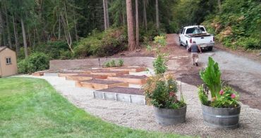 Raised garden beds and flower barrels in a forested backyard with a pickup truck.