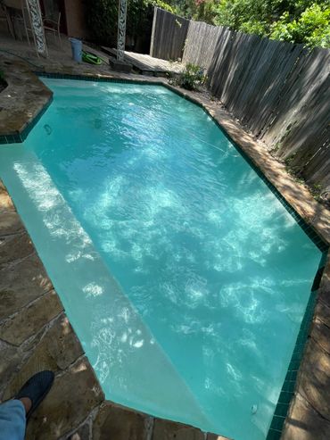 A clear backyard swimming pool surrounded by stone pavement and a wooden fence.