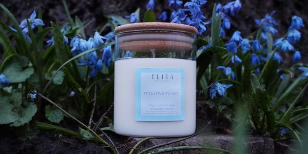 A candle labeled 'mountain rain' surrounded by blue flowers in nature.