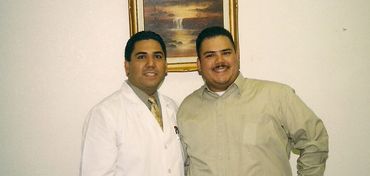Two men standing side by side indoors, one in a white lab coat, the other in casual attire.