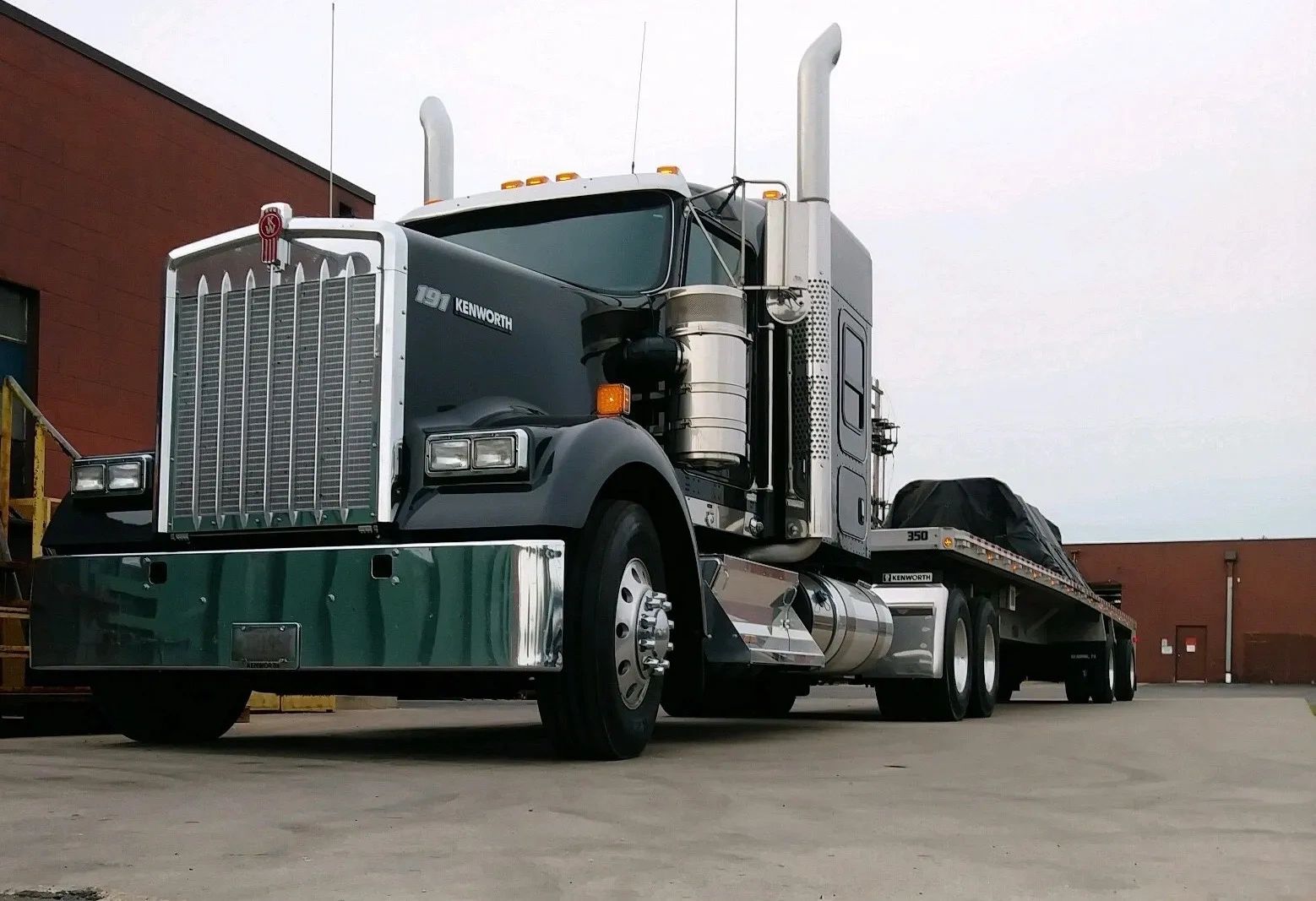 A large Kenworth semi-truck with a flatbed trailer parked near a building.