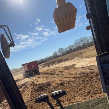 View from inside an excavator with a dirt bucket lifted and a truck in the background.