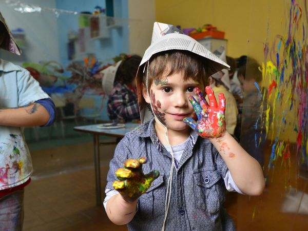 A child with painted hands and face wearing a paper hat, enjoying painting activities.