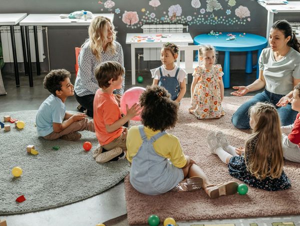 Children sitting in a circle on rugs, listening to two teachers in a classroom.