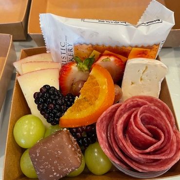 A gourmet snack box with cheese, salami rose, fruits, and crackers.
