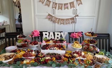 Elegant baby shower table with abundant fruits, cheeses, and charcuterie, decorated with flowers and a "Baby in Bloom" banner.