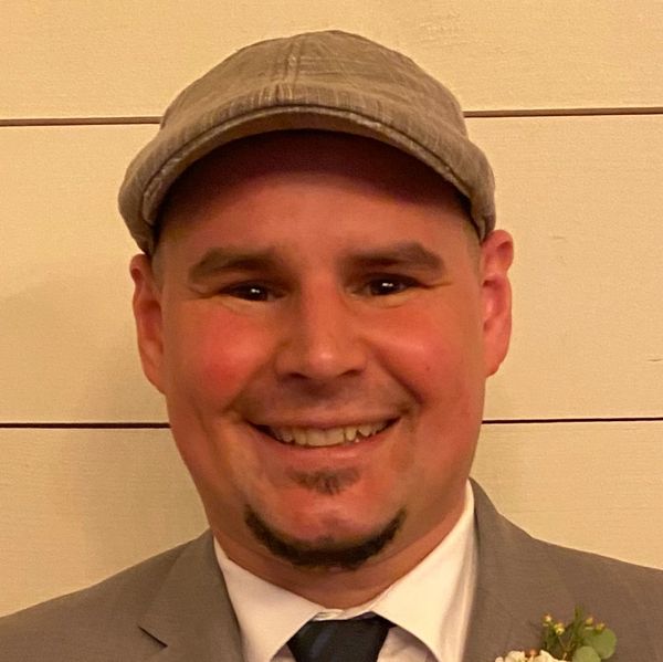 Smiling man in a gray suit and cap against a beige background.