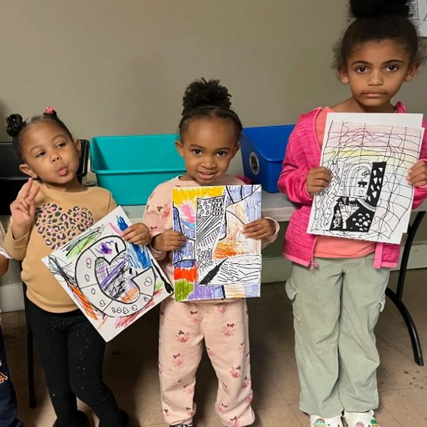 Three girls proudly display their colorful and creative drawings indoors.