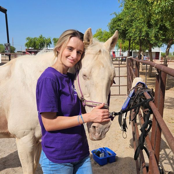 A woman with blonde hair and a purple shirt hugs a white horse