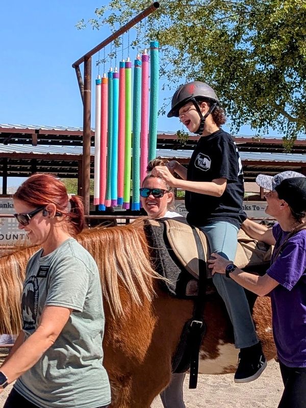 Girl with disabilities laughs while riding a horse during therapy with volunteers and a therapist.